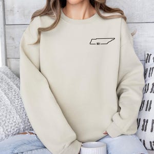 May include: Cream-colored sweatshirt with a black outline of the state of Tennessee and the number "901" printed on the upper left chest. The sweatshirt has long sleeves and a crew neck.