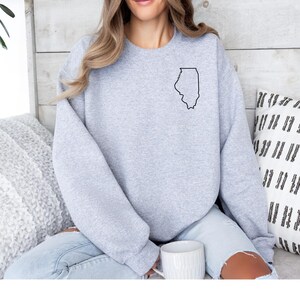 May include: A light gray sweatshirt featuring a black outline of the state of Illinois on the upper left chest. The sweatshirt is a crew neck style and is made of a soft material. The person is wearing light blue jeans and holding a white mug.
