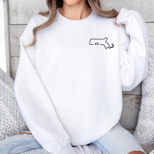 May include: A white sweatshirt with a black outline of the Massachusetts state map and the number "413" printed on the upper left chest area. The sweatshirt has long sleeves and a crew neck.