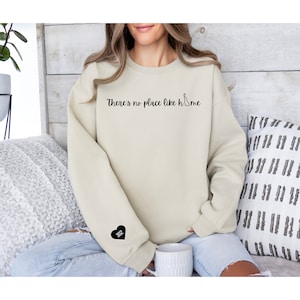May include: A beige sweatshirt with the text "There's no place like home" and a black heart with the number 302 on the left sleeve.