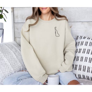May include: A beige sweatshirt with a black outline of the state of Delaware on the left chest. The outline includes the word "Delaware" written vertically inside the state.