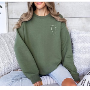 May include: Olive green sweatshirt with a white outline of the state of Vermont. The sweatshirt is a solid color and has a crew neck. The Vermont outline is on the upper left chest area. The sweatshirt is made of a soft material.