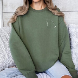 May include: Olive green sweatshirt featuring a white outline of the state of Missouri with the numbers "314" printed inside. The sweatshirt has long sleeves and a crew neck. The garment is made of a soft material.