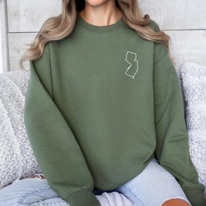 May include: Olive green sweatshirt with a white outline of the state of New Jersey on the upper chest. The sweatshirt has long sleeves and a crew neck. The person in the image is wearing light blue jeans.