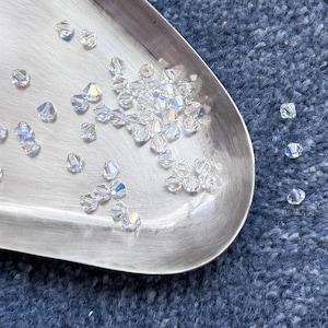 May include: Clear, faceted crystal beads are scattered on a silver metal tray. The beads have an iridescent quality, reflecting multiple colours. The tray is set against a blue textured background.