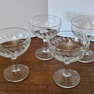May include: A set of four clear glass coupe glasses, each featuring a delicate etched laurel leaf pattern encircling the rim, complemented by two thin horizontal etched lines below the leaves, supported by a short stem and a circular base.