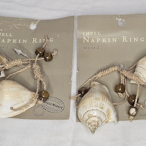 May include: Two shell napkin rings, each with two seashells, wooden beads, and a natural rope. The packaging reads "SHELL NAPKIN RING SET OF 2". The shells are cream-colored with brown accents.
