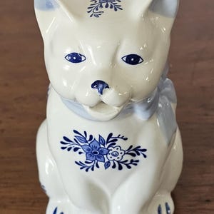 May include: A ceramic cat figurine in a sitting position, featuring a white base with blue floral patterns and accents. The cat has blue eyes, a blue bow, and blue floral designs on its head and chest.