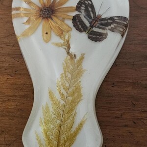 May include: A white resin spoon rest with a dried yellow flower, a fern, and a butterfly with black and white patterned wings. The spoon rest is a decorative kitchen item.