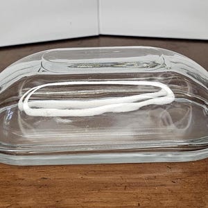 May include: A clear glass butter dish with a domed lid. The dish is oval-shaped with a slightly raised base and a matching lid. The glass is transparent, allowing the contents to be visible. The dish is designed for storing and serving butter.