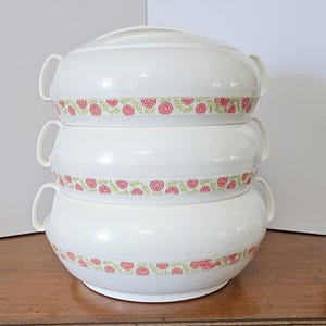 May include: A stack of three white serving bowls with lids and handles. Each bowl has a decorative band of red floral and green leaf pattern around the middle. The bowls are stacked on top of each other.