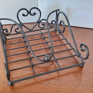 May include: A wrought iron book stand with a decorative heart and scroll design. The black metal stand has a rectangular base with a sloped back and curved supports. The stand is designed to hold books or display items.