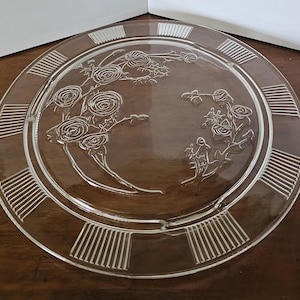 May include: A clear glass plate with an embossed floral design featuring roses. The plate has a decorative border with vertical lines. The plate is round and appears to be vintage.