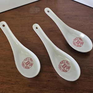May include: Three white ceramic soup spoons with a red emblem in the bowl and gold trim. Each spoon has a small hole at the end of the handle. The spoons are arranged on a dark wooden surface.