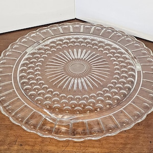 May include: A clear glass serving plate with a raised, textured design. The plate has a scalloped edge and a central design of a sunburst with a circle in the center.