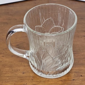 May include: A clear glass mug with a floral design etched into the side. The mug has a handle and is perfect for enjoying a hot beverage.