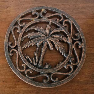May include: A circular metal trivet featuring a central palm tree motif surrounded by an intricate scrollwork border. The object has a dark bronze-toned finish with lighter copper-colored highlights on the raised surfaces of the design.