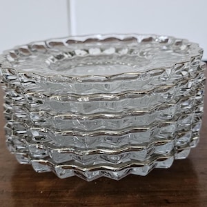 May include: A stack of clear glass coasters with a silver trim. The coasters have a textured, ridged design around the edges. The stack is on a wooden surface.