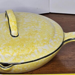 May include: Yellow ceramic casserole dish with a lid and handle. The dish has a speckled pattern and a black trim. The handle is arched and black-lined. The pan has a long handle. A kitchenware item.