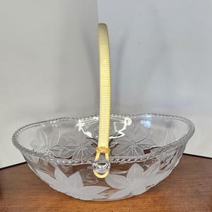 May include: A clear glass basket with a floral design and a yellow handle. The basket has a scalloped edge and is likely used for serving or display. The handle is made of a woven material.