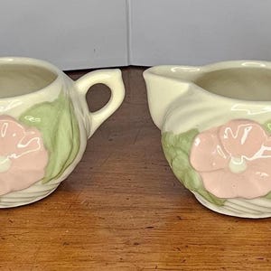 May include: A cream-colored ceramic sugar bowl and creamer set. Each piece features a pink flower with green leaves and a handle. The sugar bowl has two handles, while the creamer has a spout.