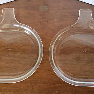 May include: Two clear glass Pyrex casserole dish lids with a handle. The lids are oval-shaped with a rounded handle at one end. The glass is transparent, and the lids are resting on a wooden surface.