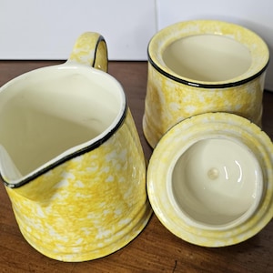 May include: A yellow and white ceramic creamer and sugar bowl set. The creamer has a handle and spout, and the sugar bowl has a lid. Both pieces have a black trim around the rim. The set has a textured, speckled design.