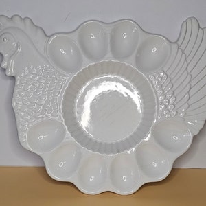 May include: White ceramic egg dish shaped like a hen with a raised wing and a head with a beak. The dish has eight individual egg-shaped indentations.