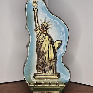 May include: A decorative bottle shaped like the Statue of Liberty. The bottle is gold with a blue and green background. The bottle has a black cap and the text "BEAM 100" and "Kentucky Straight Bourbon Whiskey".