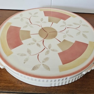 May include: A round, white ceramic cake stand with a decorative design. The top features a pattern of pink, tan, and yellow geometric shapes and leaf-like details. The rim and base are white with a scalloped edge.