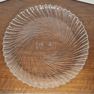 May include: A clear glass plate with a spiral design. The plate has a textured surface with a series of ridges that radiate from the center to the edge. The plate is round and has a scalloped edge.