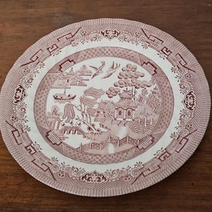 May include: A round, white and maroon plate with a detailed Willow pattern design. The plate features a central scene with a pagoda, bridge, and boat, surrounded by a decorative border. The plate is made of ceramic.