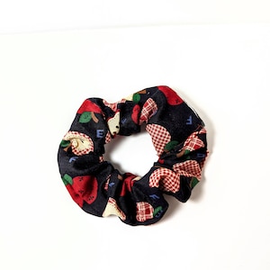 May include: A black scrunchie with a red and white apple print. The apples have a green leaf and a red stem.
