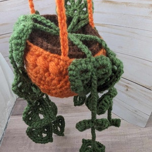 May include: A crocheted pumpkin-shaped hanging planter with green leaves. The planter is orange and brown and has a hanging loop.