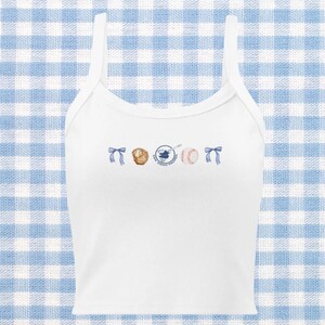Pode incluir: Camisola branca de alças com um design de temática de beisebol. O design apresenta uma luva de beisebol, uma bola de beisebol, um taco e uma fita azul.