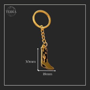 May include: Gold-tone metal cowboy boot keychain with a black enamel finish. The boot features a cactus and star design. The keychain measures 30mm in height and 18mm in width.