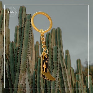 May include: Gold-tone keychain featuring a black and green cowboy boot with a cactus and star design. The keychain is hanging from a cactus plant.