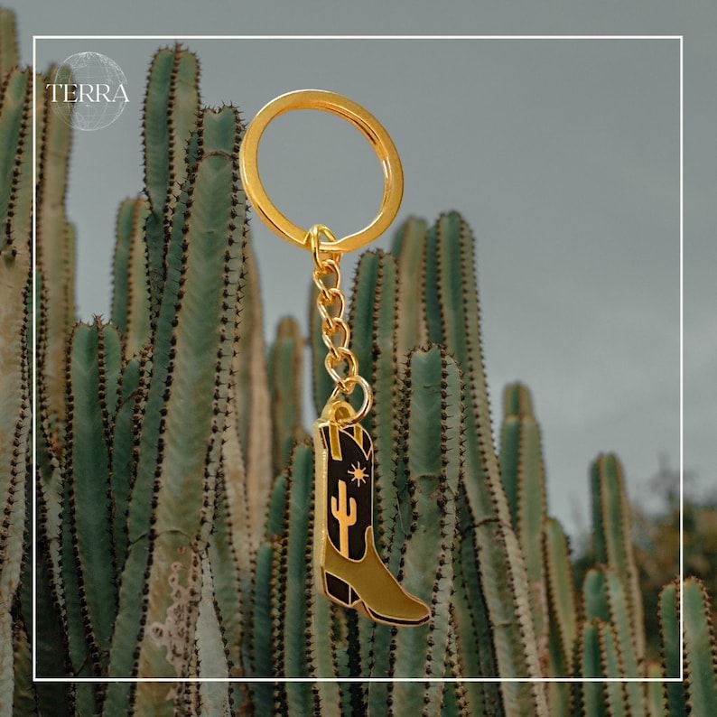 May include: Gold-tone keychain featuring a black and green cowboy boot with a cactus and star design. The keychain is hanging from a cactus plant.