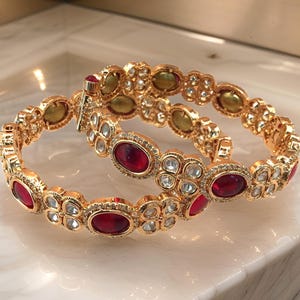 May include: Two gold bangles with red and white stones. The bangles are decorated with intricate floral designs.