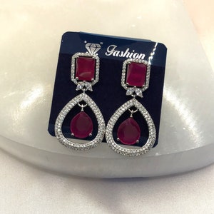 May include: A pair of silver dangle earrings with a red square stone and a teardrop-shaped red stone. The earrings are on a blue card with the word "Fashion" printed on it.