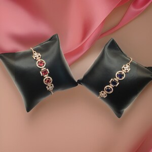 May include: Two gold bracelets with square-shaped gemstones. One bracelet has red gemstones and the other has blue gemstones. Both bracelets are displayed on black velvet cushions.