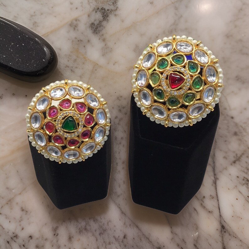 Puede incluir: Dos piezas de joyer&iacute;a redondas de color dorado con detalles de perlas blancas. Cada pieza presenta una piedra preciosa central verde y roja rodeada de piedras preciosas verdes y rojas m&aacute;s peque&ntilde;as.
