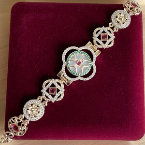 May include: A gold bracelet with a white and green floral design and red gemstones. The bracelet is on a red velvet background.