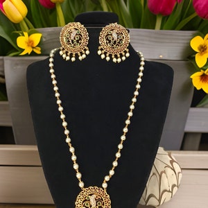 May include: A gold necklace with a round pendant featuring a green and red floral design. The pendant is surrounded by white pearls. The necklace is displayed on a black stand. The necklace comes with a matching pair of earrings with the same design.