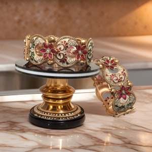 May include: Two gold bangles with intricate floral designs and red and white enamel. The bangles are decorated with small, sparkling stones.