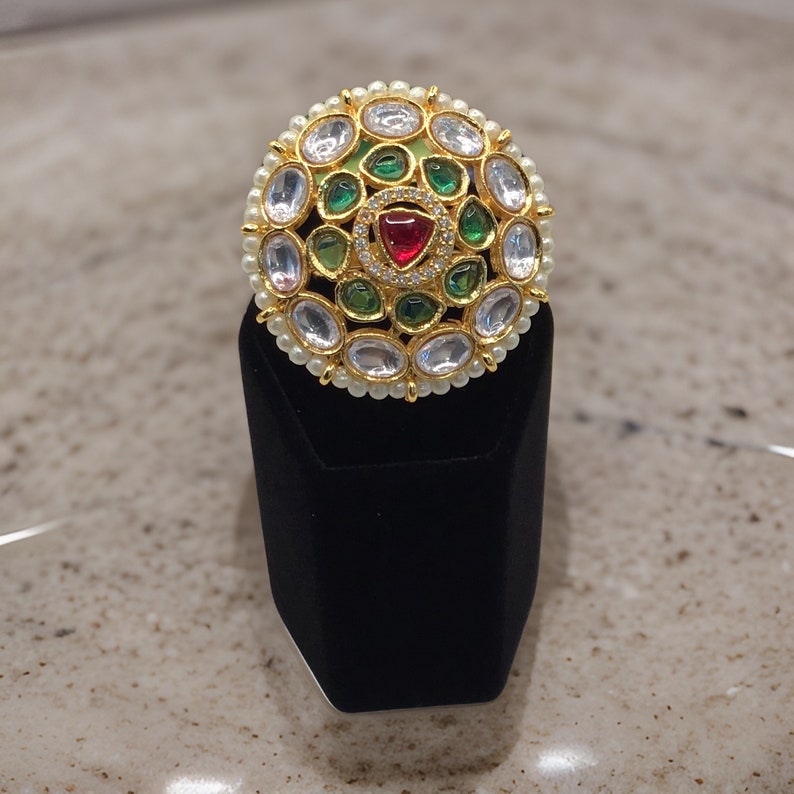 Puede incluir: Un anillo de oro con un dise&ntilde;o redondo con piedras preciosas blancas, verdes y rojas. El anillo est&aacute; colocado en un expositor de anillos de terciopelo negro.