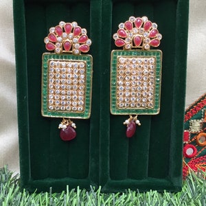May include: A pair of gold earrings with green and red stones. The earrings are rectangular with a design of small white stones. The earrings are in a green velvet box.