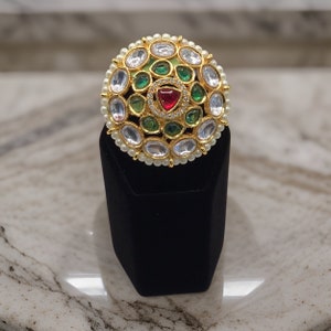 Puede incluir: Un anillo de oro con un dise&ntilde;o redondo con piedras preciosas blancas, verdes y rojas. El anillo est&aacute; colocado en un expositor de terciopelo negro.