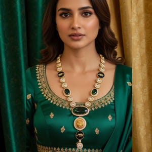 May include: A woman wearing a green satin blouse with gold embroidery and a gold necklace with emerald and black accents. The necklace features a large oval emerald pendant and a lion emblem.