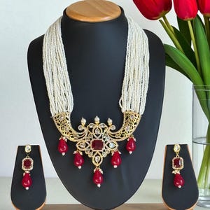 May include: An ornate necklace and earring set displayed on a black mannequin. The necklace features a gold-toned filigree pendant with a large red square stone and smaller red teardrop-shaped stones. The earrings mirror the necklace design.